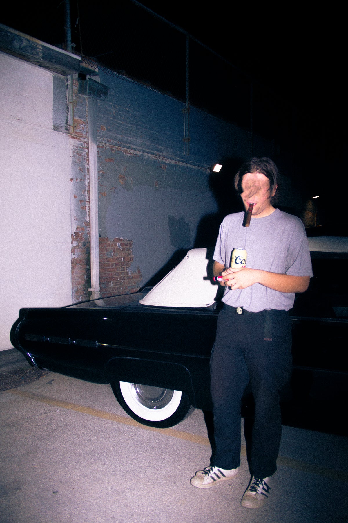 A person standing outdoors at night beside a black vintage car, wearing a light gray t-shirt, dark pants, and sneakers while holding a drink. The photo has a grainy, low-light aesthetic.