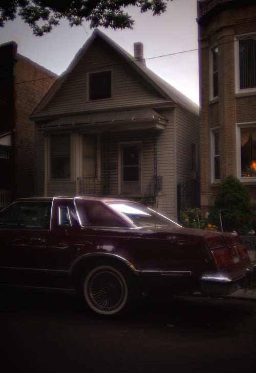 A dark maroon vintage Cadillac sedan parked on a residential street in front of a gray house.