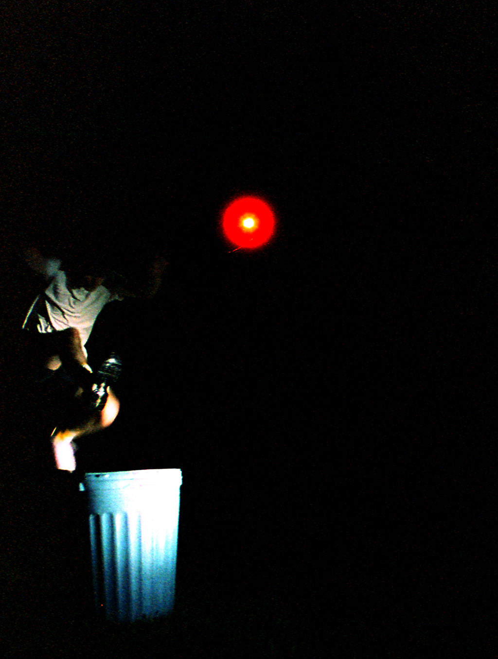 A person in a white shirt jumps onto a white trash can in a very dark outdoor setting, with a bright red light or reflector shining in the air above them.