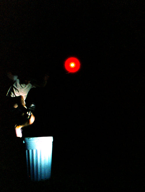 A person in a white shirt jumps onto a white trash can in a very dark outdoor setting, with a bright red light or reflector shining in the air above them.