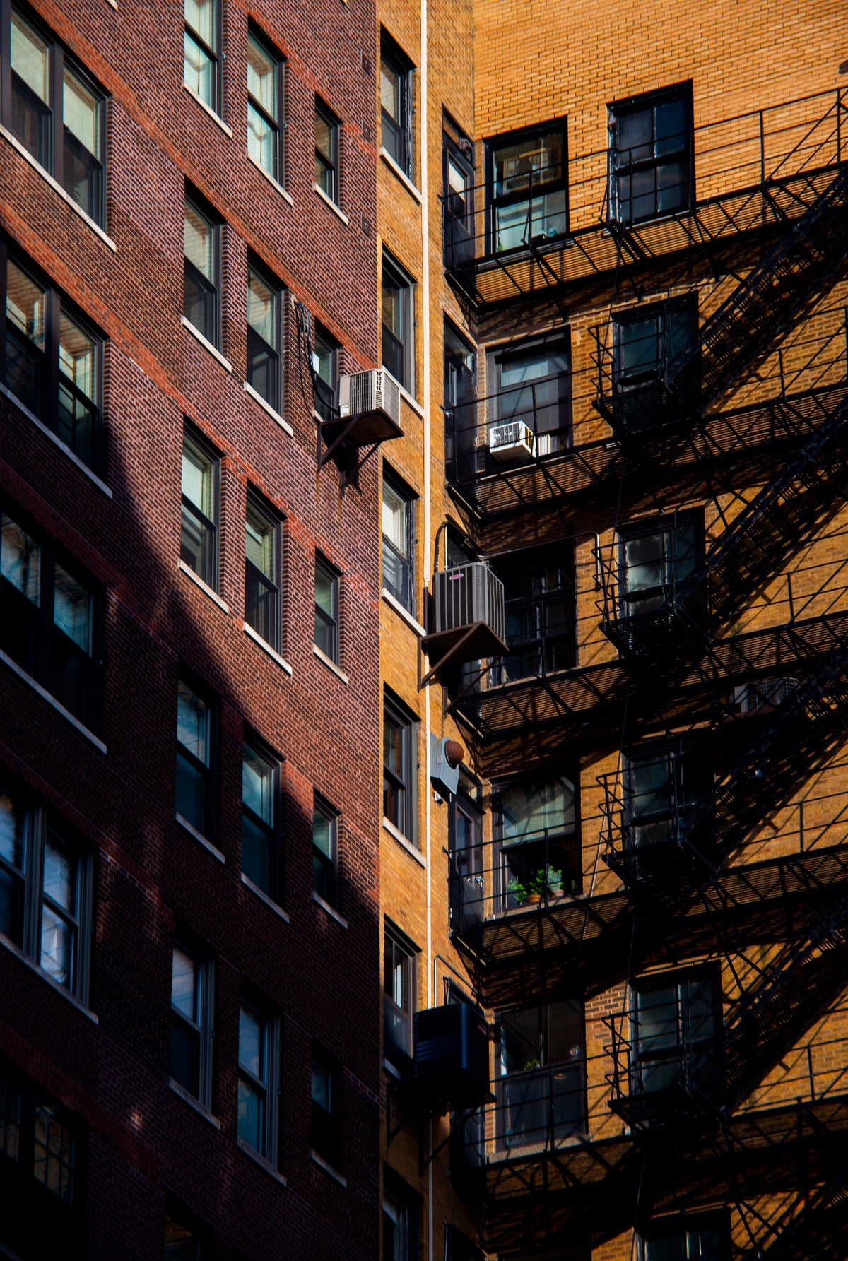A low-angle photograph of two tall brick apartment buildings, one in dark red-brown brick and the other in warm orange-brown brick, with black fire escapes on the right side casting strong shadows.