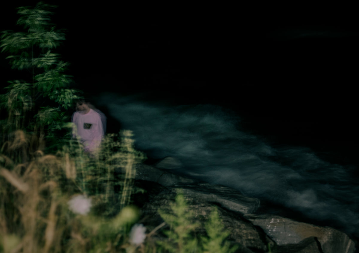 A person in a light pink hoodie stands on a rocky riverbank at night, with dark water and blurred green foliage in the background.
