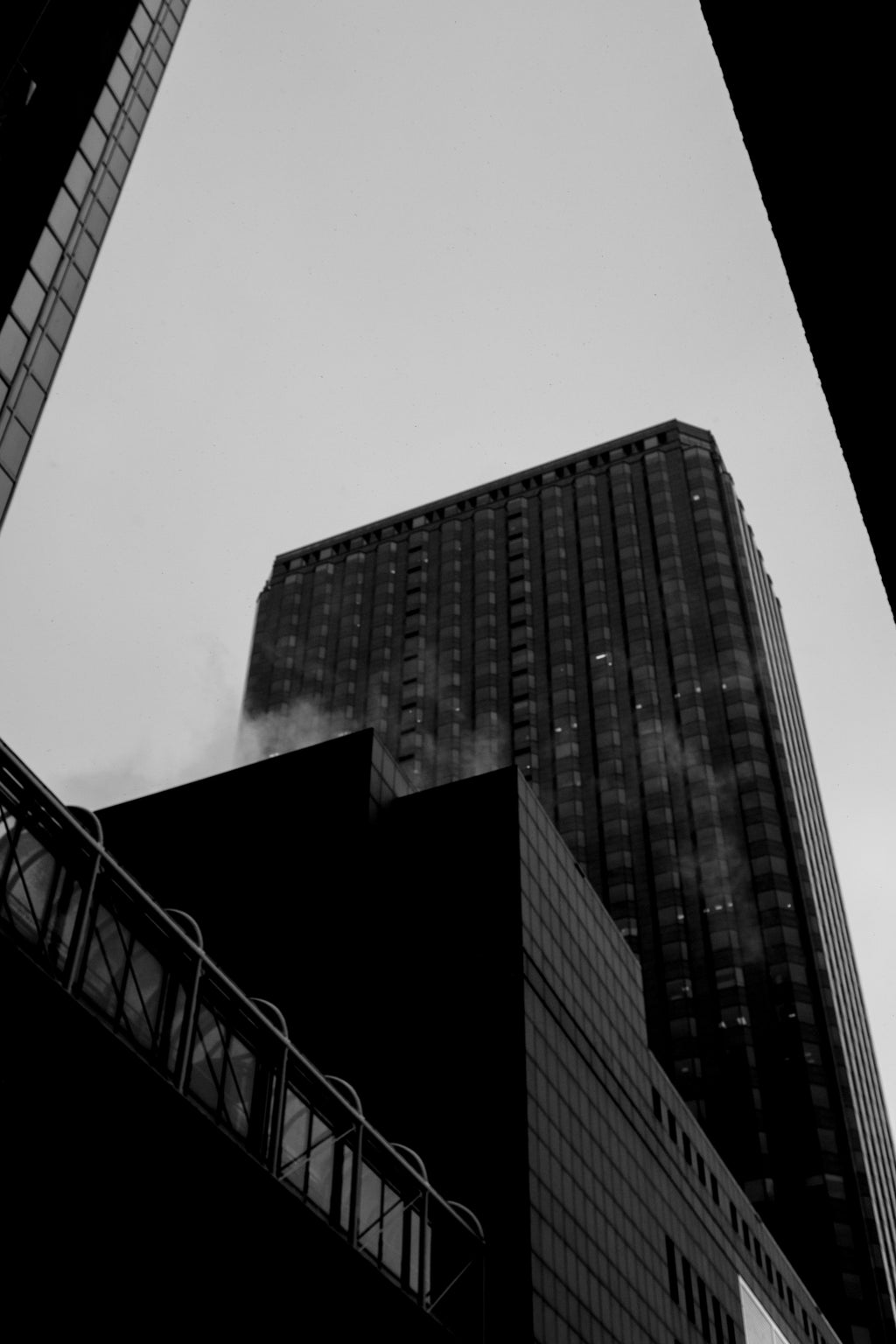 Black and white architectural photograph of a tall modern skyscraper viewed from below, framed by surrounding buildings against a pale sky.