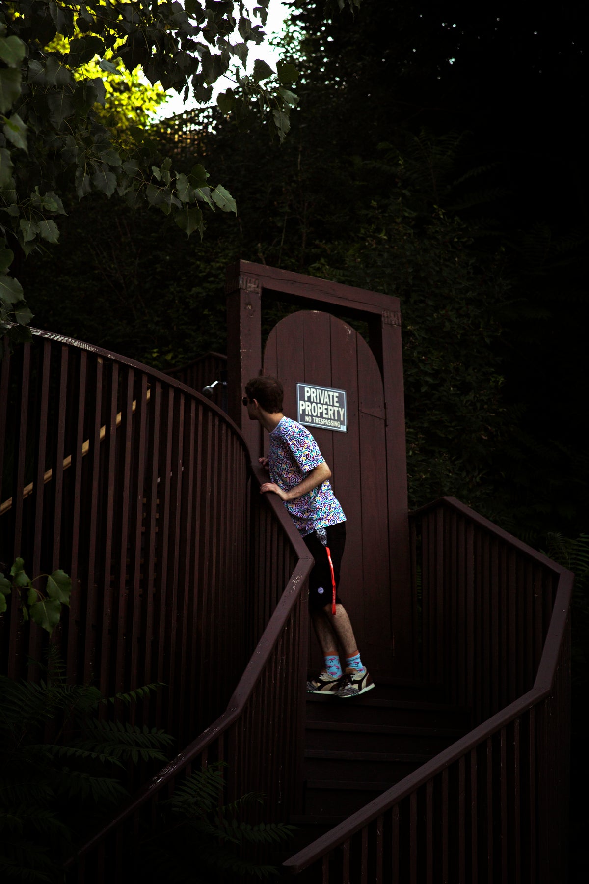 A person in athletic wear stands on dark wooden stairs in a forest setting, facing a small arched door with a sign that reads 'PRIVATE PROPERTY'.