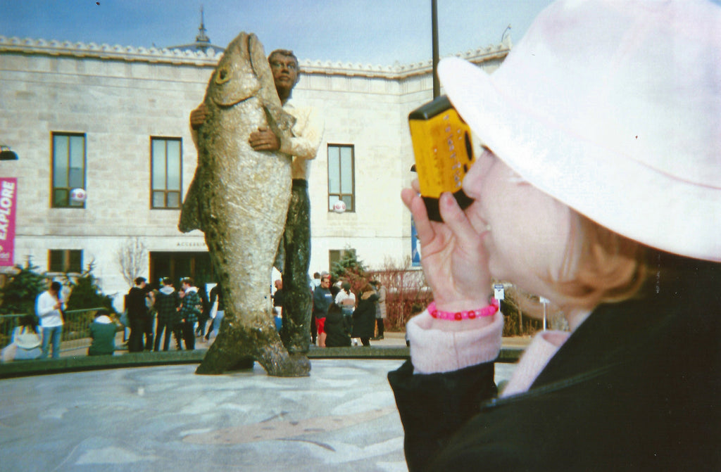 A woman in a white sun hat and dark coat holds a yellow walkie-talkie near her face, standing outdoors in front of a large stone building and a fish statue. Other people are visible in the background.