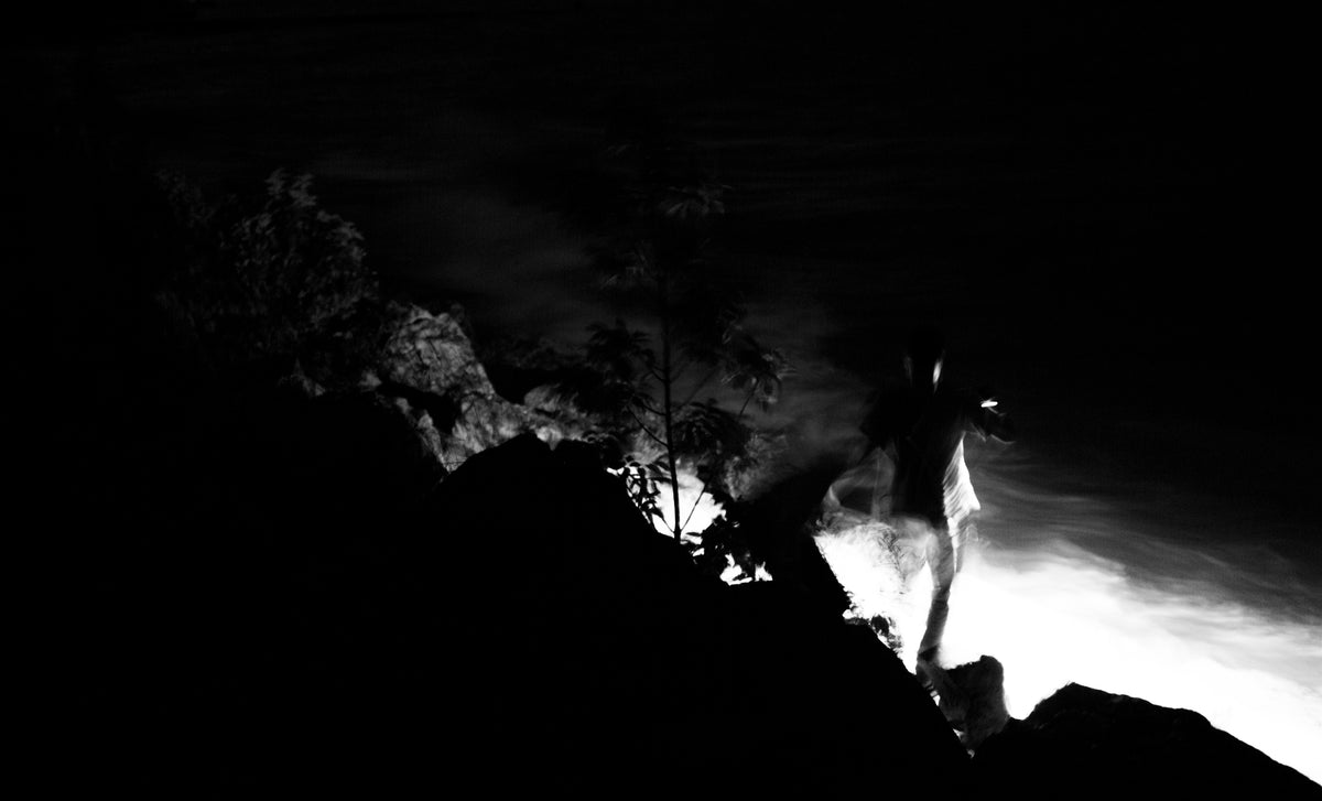 Black and white dramatic photograph of a man standing on a rocky shoreline at night, with bright light reflecting off the water and dark silhouetted rocks and trees.