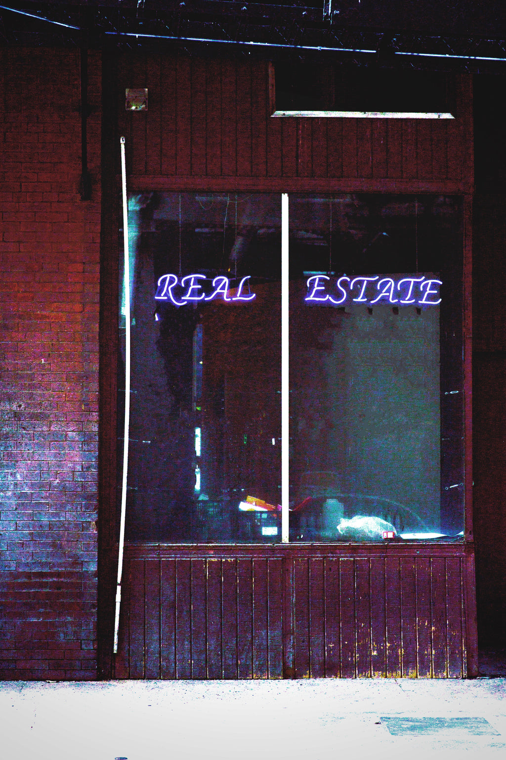 A storefront window with a dark brick wall and a purple neon sign reading 'REAL ESTATE' across the glass.
