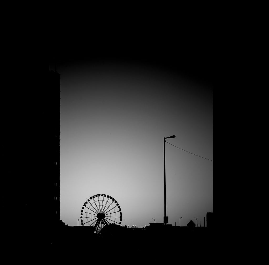 Black and white square art print featuring the silhouette of a ferris wheel and a street lamp against a gradient gray sky framed by dark building silhouettes.