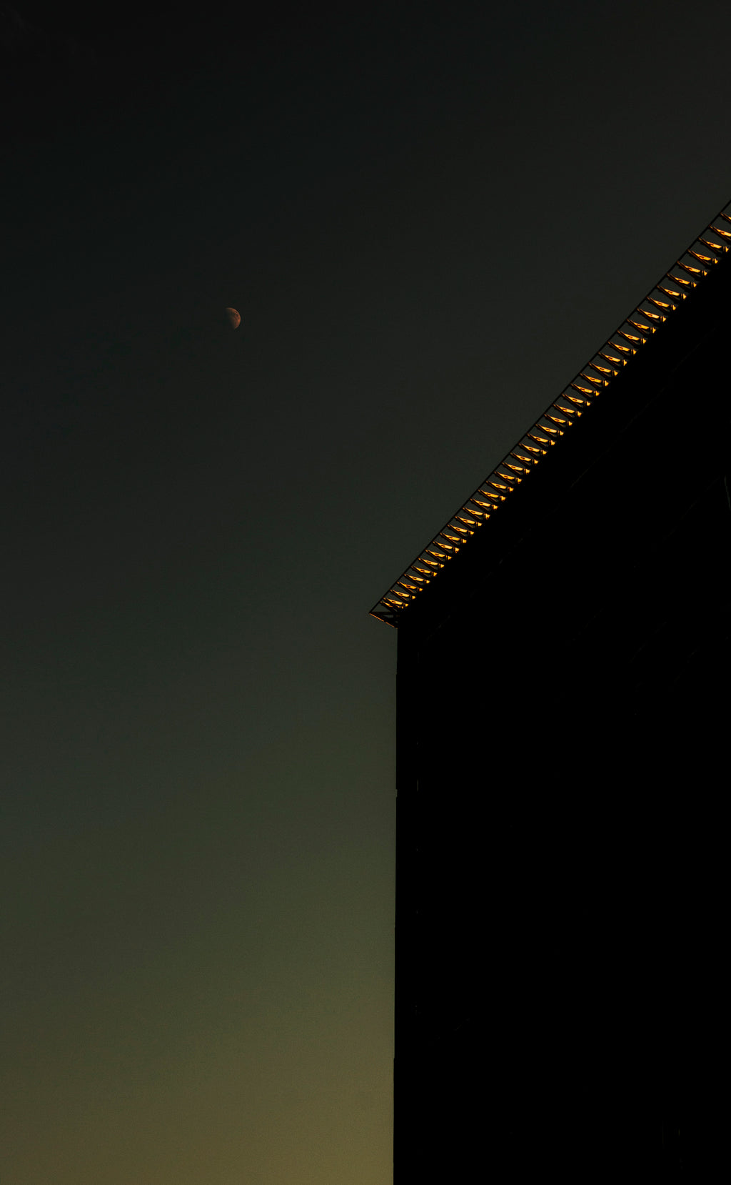 A dark architectural silhouette of a building corner with warm glowing lights along the roofline, set against a twilight sky with a visible crescent moon.