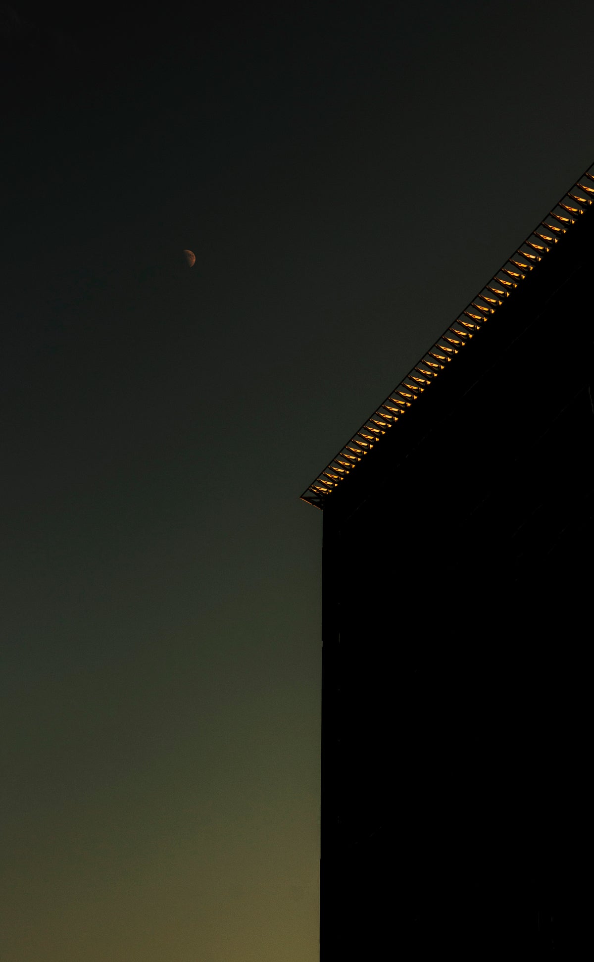 A dark architectural silhouette of a building corner with warm glowing lights along the roofline, set against a twilight sky with a visible crescent moon.