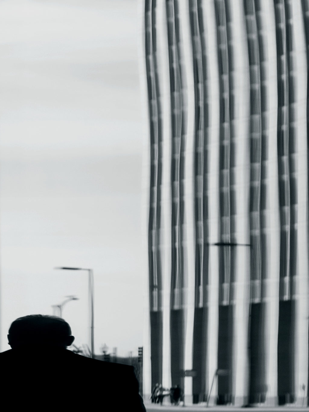 Black and white street photograph showing the silhouette of a person on the left looking toward a tall modern building with vertical window lines on the right.