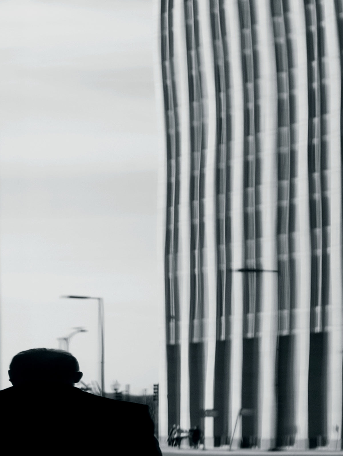 Black and white street photograph showing the silhouette of a person on the left looking toward a tall modern building with vertical window lines on the right.